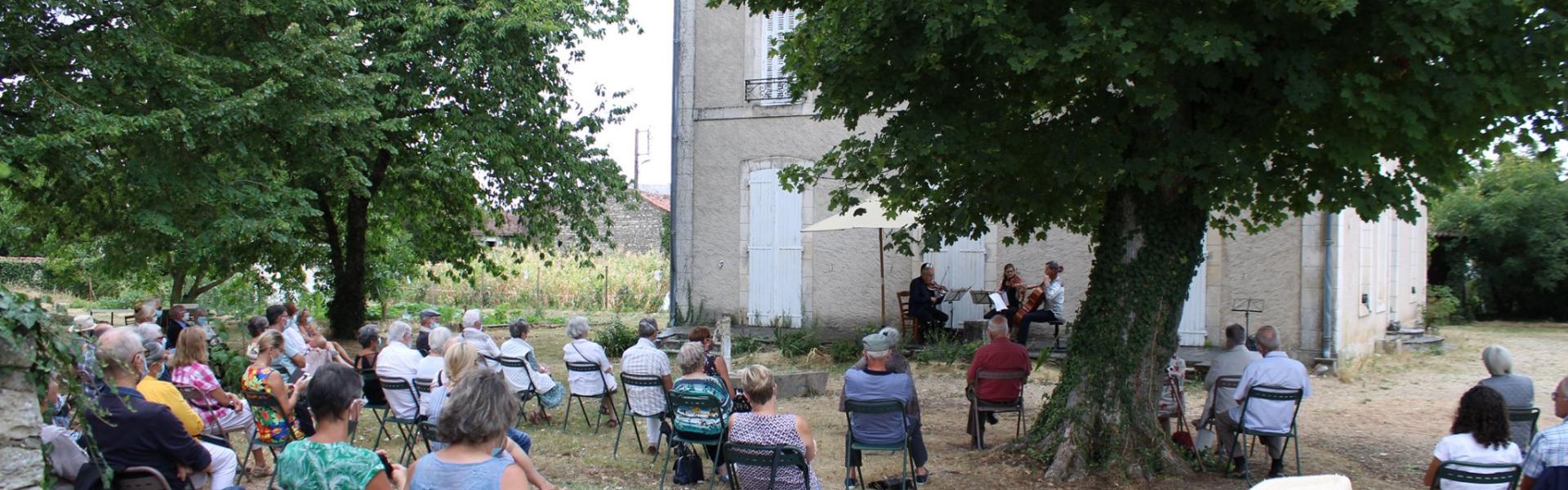 Une photo d'un concert à la Maison Tassin. Du public assis en plein air écoute des musiciens.