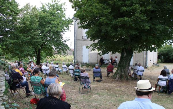 Une photo d'un concert à la Maison Tassin. Du public assis en plein air écoute des musiciens.