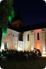 Une photo de nuit à l'extérieur du Prieuré Saint-André éclairé. Le public écoute les musiciens à côté d'un palmier.